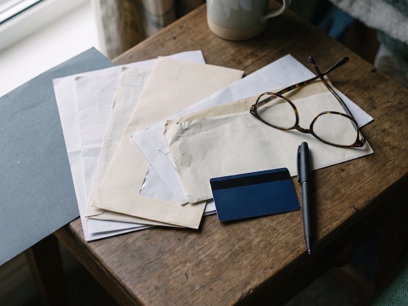 A pile of unopened letters, a bank card, reading glasses and a pen on a wooden table by a window
