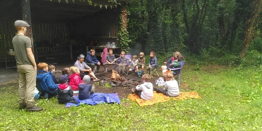 Group of children sitting on grass around a campfire at the edge of a forest,